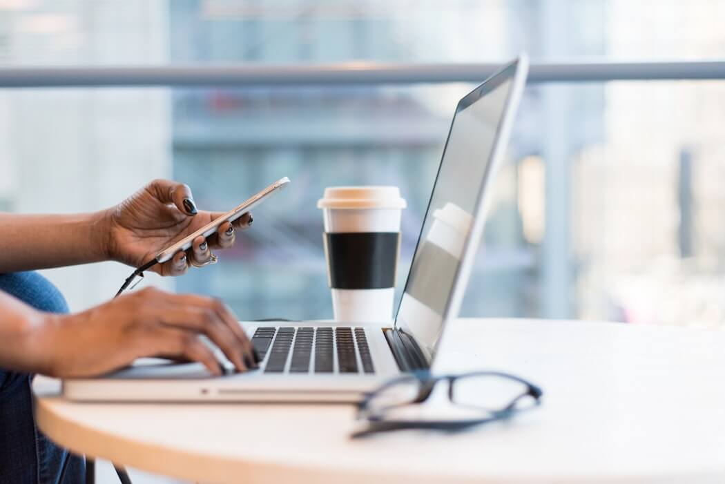 A person uses a smartphone and laptop at a round table with a takeaway coffee cup and eyeglasses, with large windows and a blurred cityscape in the background, suggesting a modern, productive workspace, ideal for a social media scheduler | plannthat.com