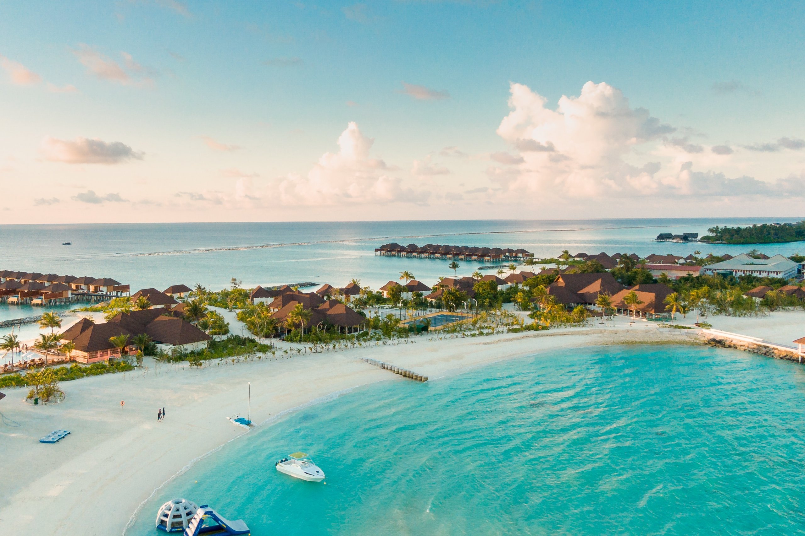 Aerial view of a tropical beach resort with overwater bungalows, turquoise waters, white sandy beaches, and lush greenery under a clear blue sky with scattered clouds. A boat is anchored near the shore, and a few people are walking, perfect for sharing on Instagram using a social media scheduler | plannthat.com