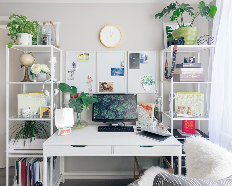 A well-organized home office with a white desk, computer, plants, and decorative items. Shelves hold books, a globe, and various decor. A clock hangs on the wall above the desk, creating a clean and inspiring workspace, perfect for using a social media planner. | plannthat.com