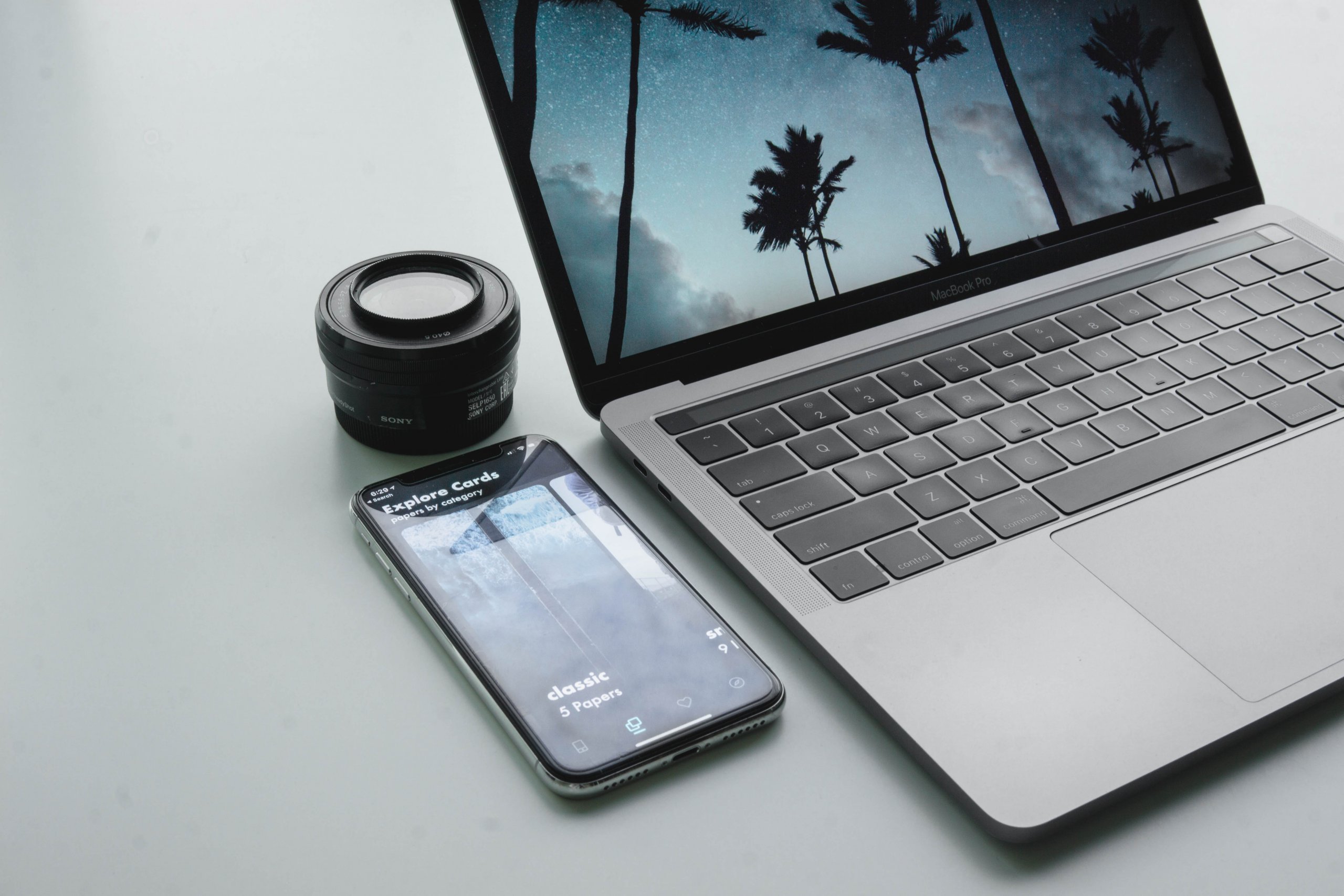 A laptop displaying a tropical palm tree wallpaper, a smartphone showing an app, and a camera lens on a white surface, suggesting a workspace setup for content creation or personal branding with a social media scheduler | plannthat.com