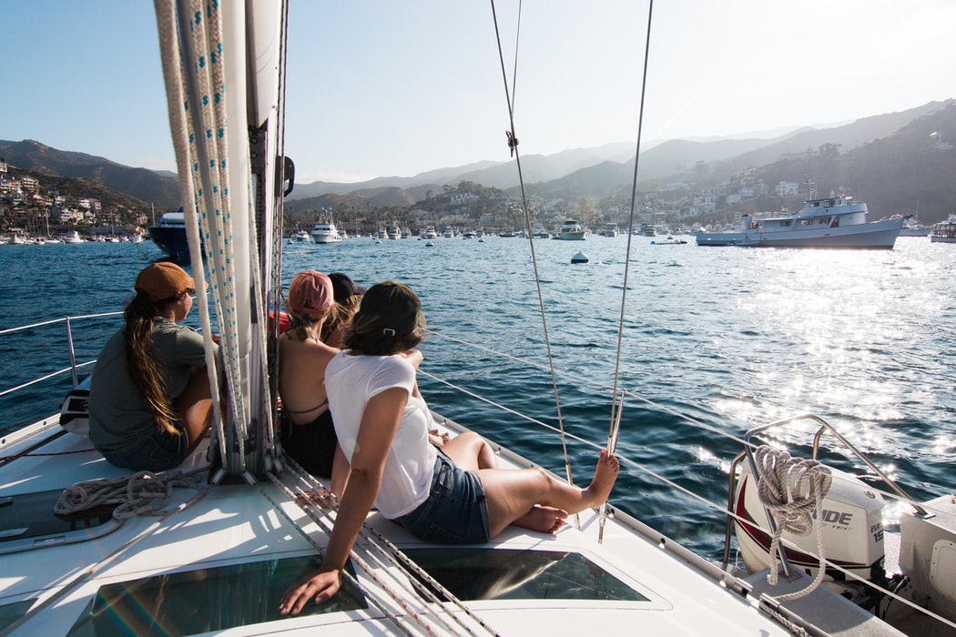 People relaxing on a sailboat, enjoying the view of a harbor filled with boats, with mountains and a coastal town in the background, perfect for sharing with a social media scheduler | plannthat.com