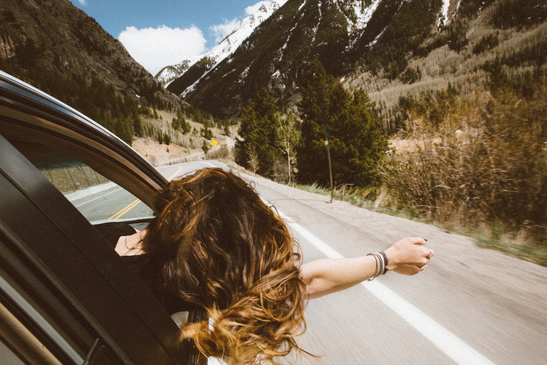 A person with long hair leans out of a car window, enjoying a scenic drive through a mountainous landscape with clear skies and lush greenery, perfect for capturing moments with an Instagram scheduler | plannthat.com
