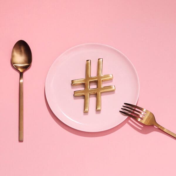 A gold hashtag symbol on a pink plate, with a gold spoon on the left and a gold fork on the right, against a pink background, representing a social media planner | plannthat.com