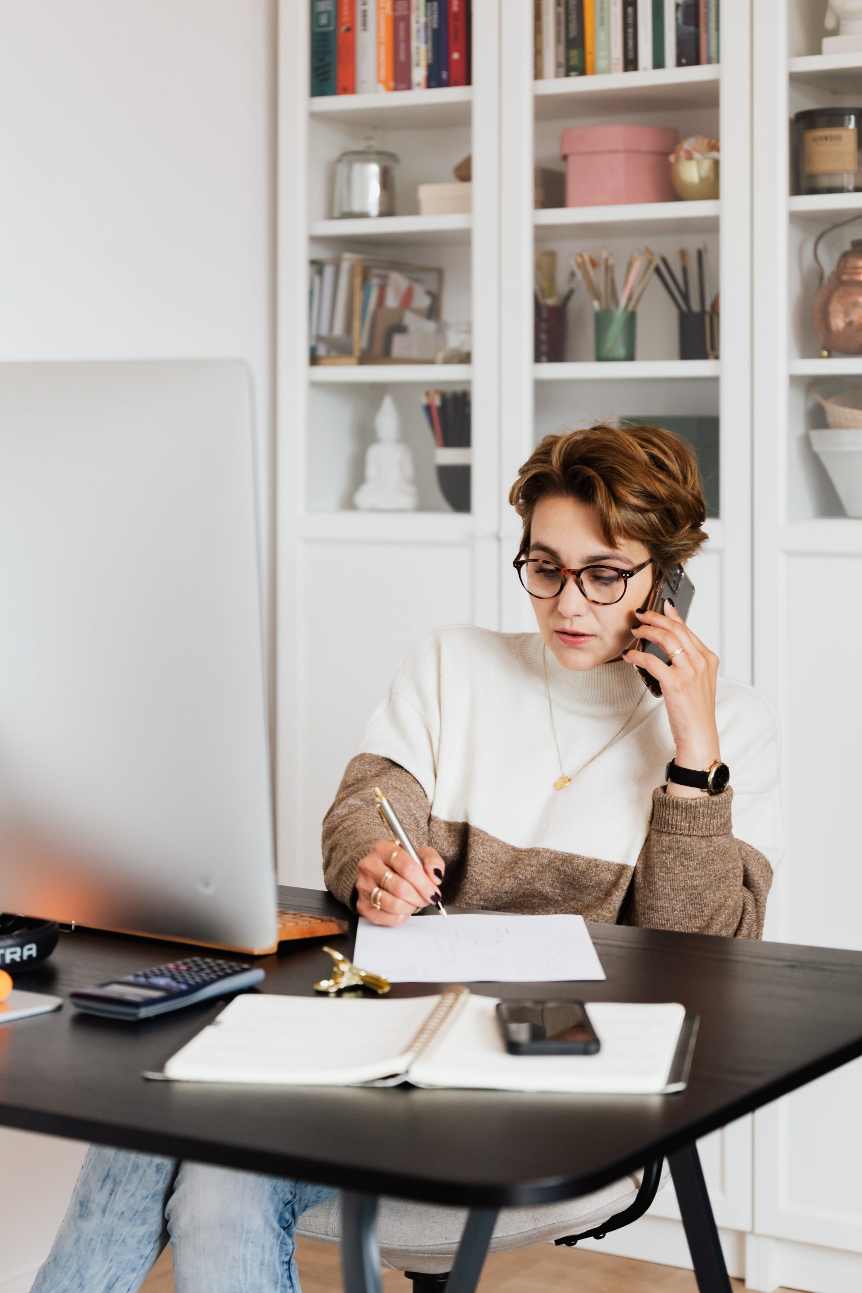 Woman working at a desk, talking on the phone, and writing notes with a computer, notebook, and calculator on the table in a home office with shelves in the background, using a social media scheduler | plannthat.com