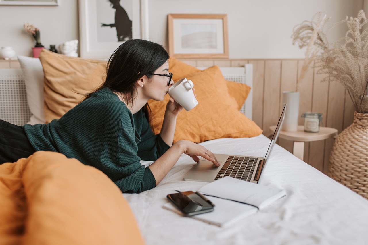 Woman working on a laptop in bed, drinking coffee, with a notepad and phone beside her, surrounded by orange pillows and cozy decor | Social media planner | plannthat.com