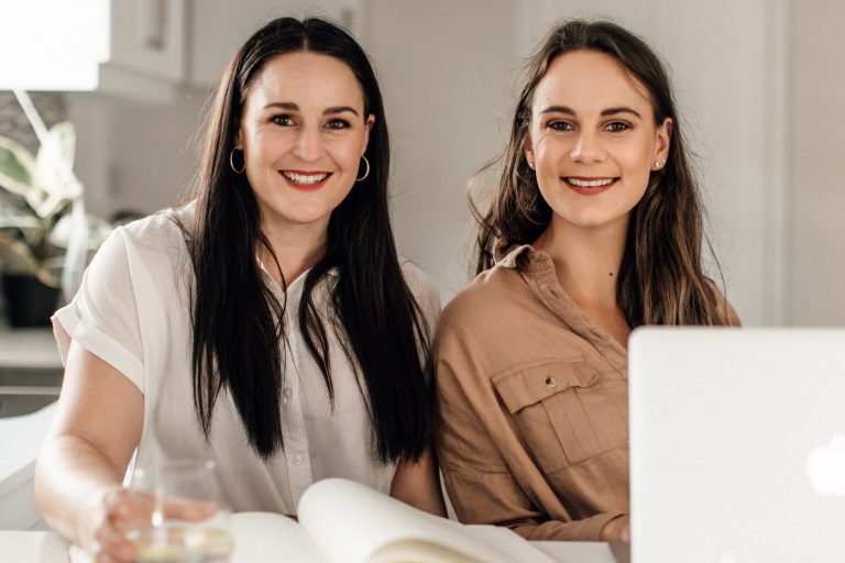 Two women smiling, seated at a table with a laptop and open book in a bright indoor setting, using a social media scheduler | plannthat.com