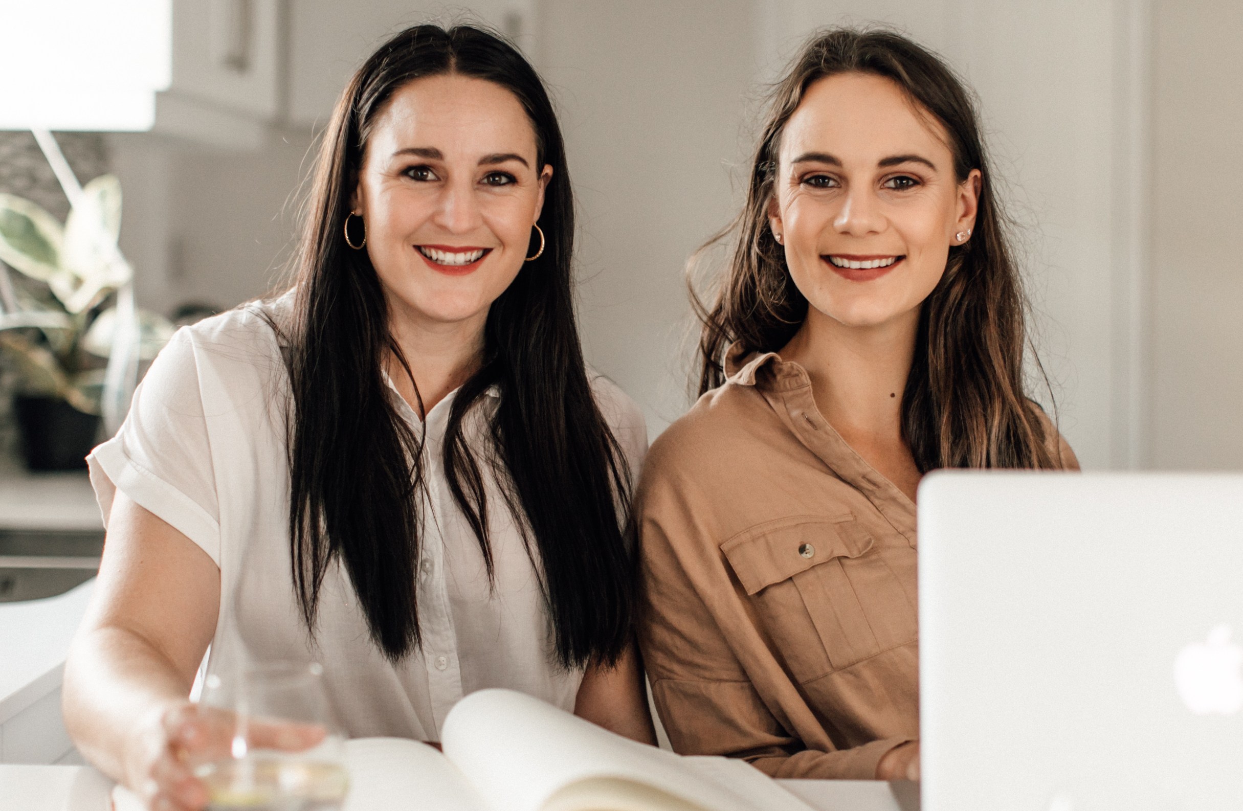 Two women smiling, seated at a table with a laptop and open book in a bright indoor setting, using a social media scheduler | plannthat.com