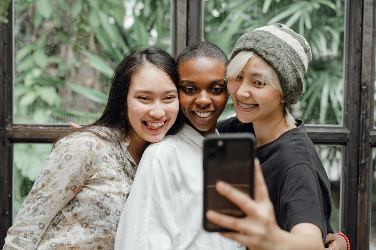 Three friends smiling and taking a selfie together indoors, with lush green plants visible through large windows behind them, appearing happy and relaxed in a bright, natural setting, perfect for a social media planner | plannthat.com