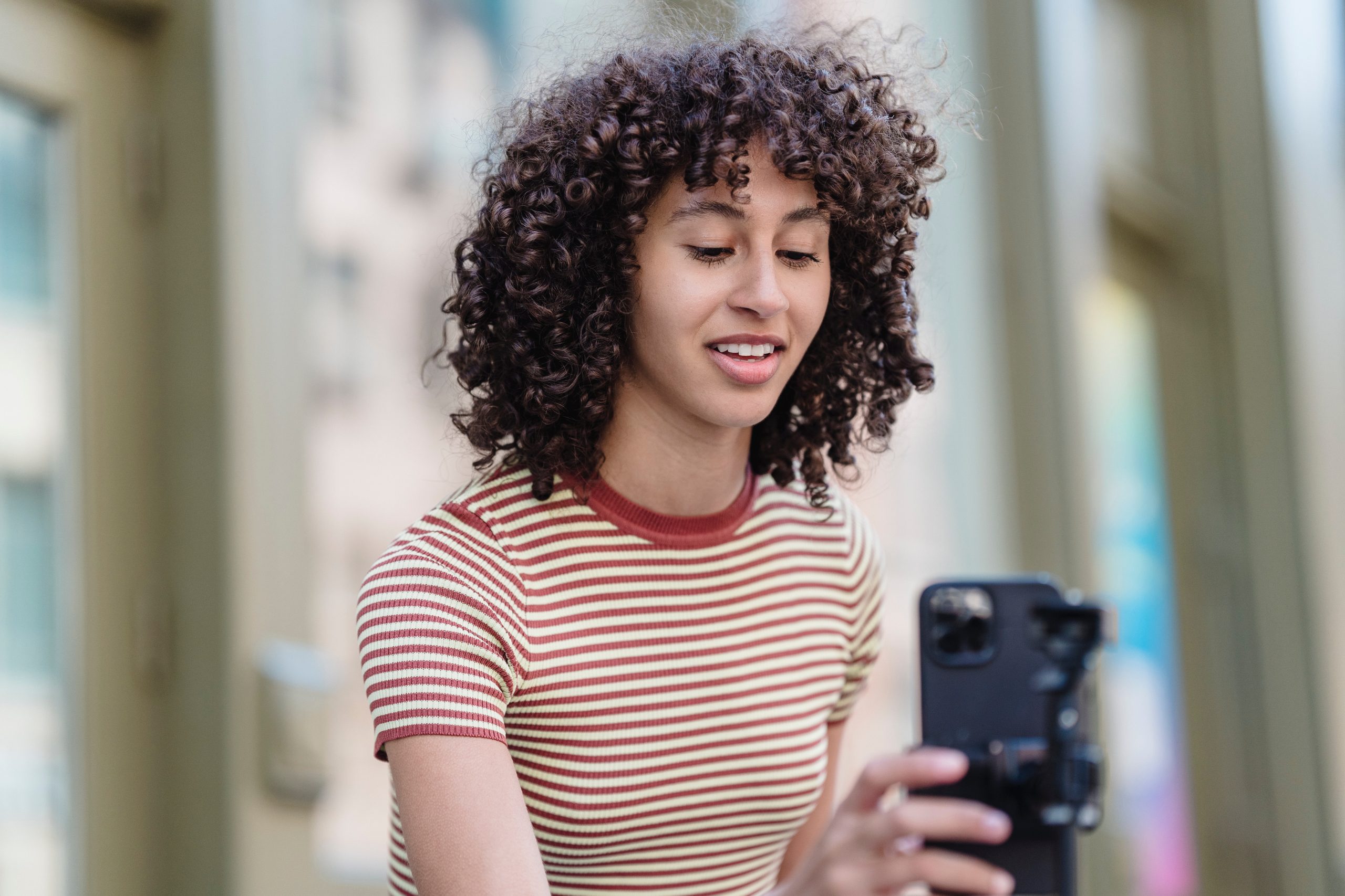 Person with curly hair wearing a striped shirt, holding a smartphone and smiling outdoors, using a social media scheduler | plannthat.com