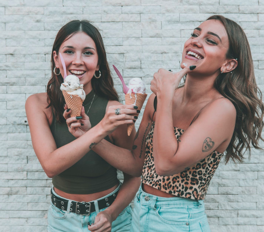 Two women smiling and holding ice cream cones in front of a light brick wall, one in a green top and the other in a leopard print top, both appearing happy and enjoying their ice cream | Social media scheduler | plannthat.com