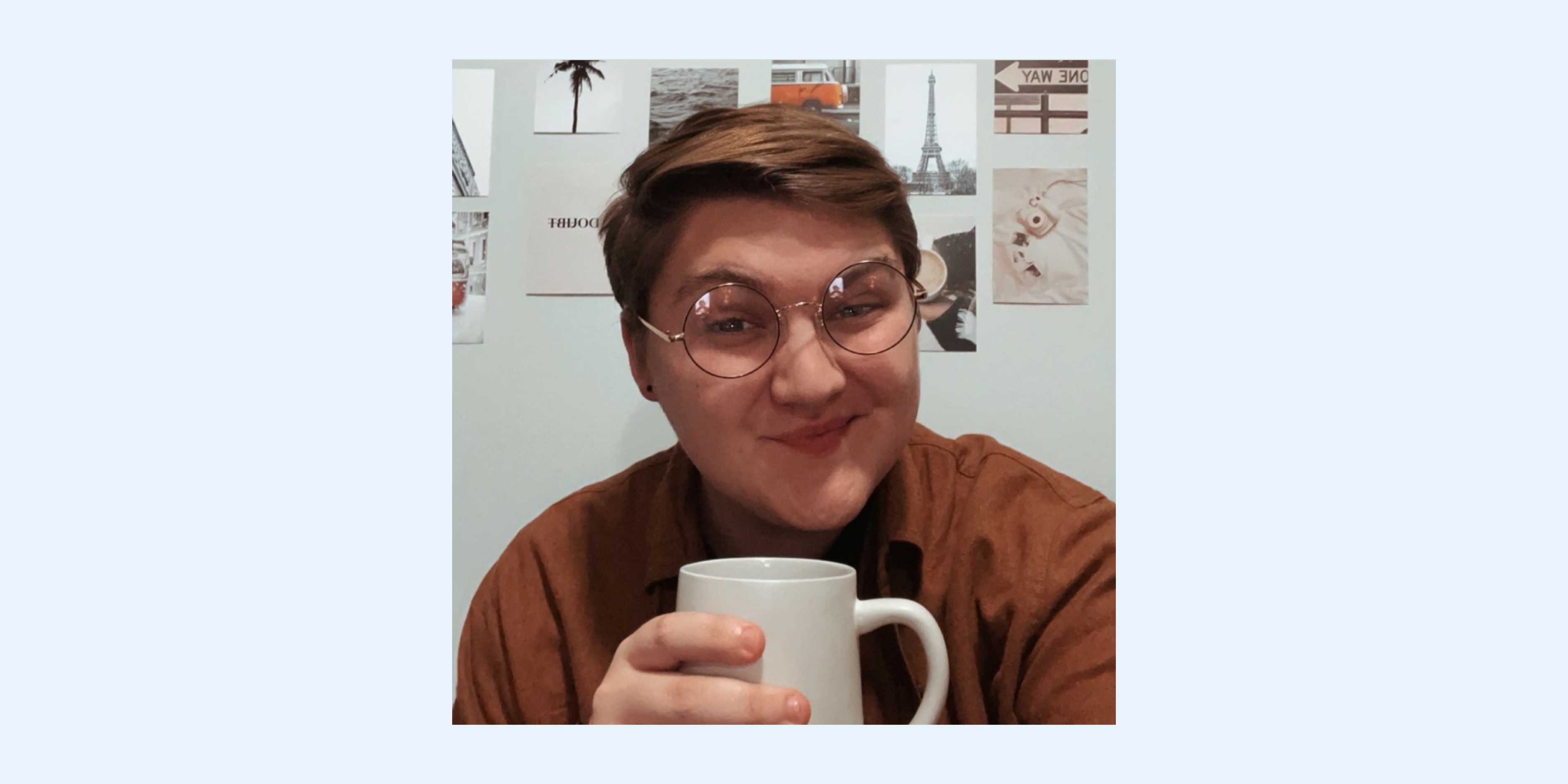 A person with short hair and glasses smiles while holding a white mug, with travel-themed posters of the Eiffel Tower and a palm tree in the background, creating a cozy and inspiring atmosphere, perfect for using a social media scheduler | plannthat.com