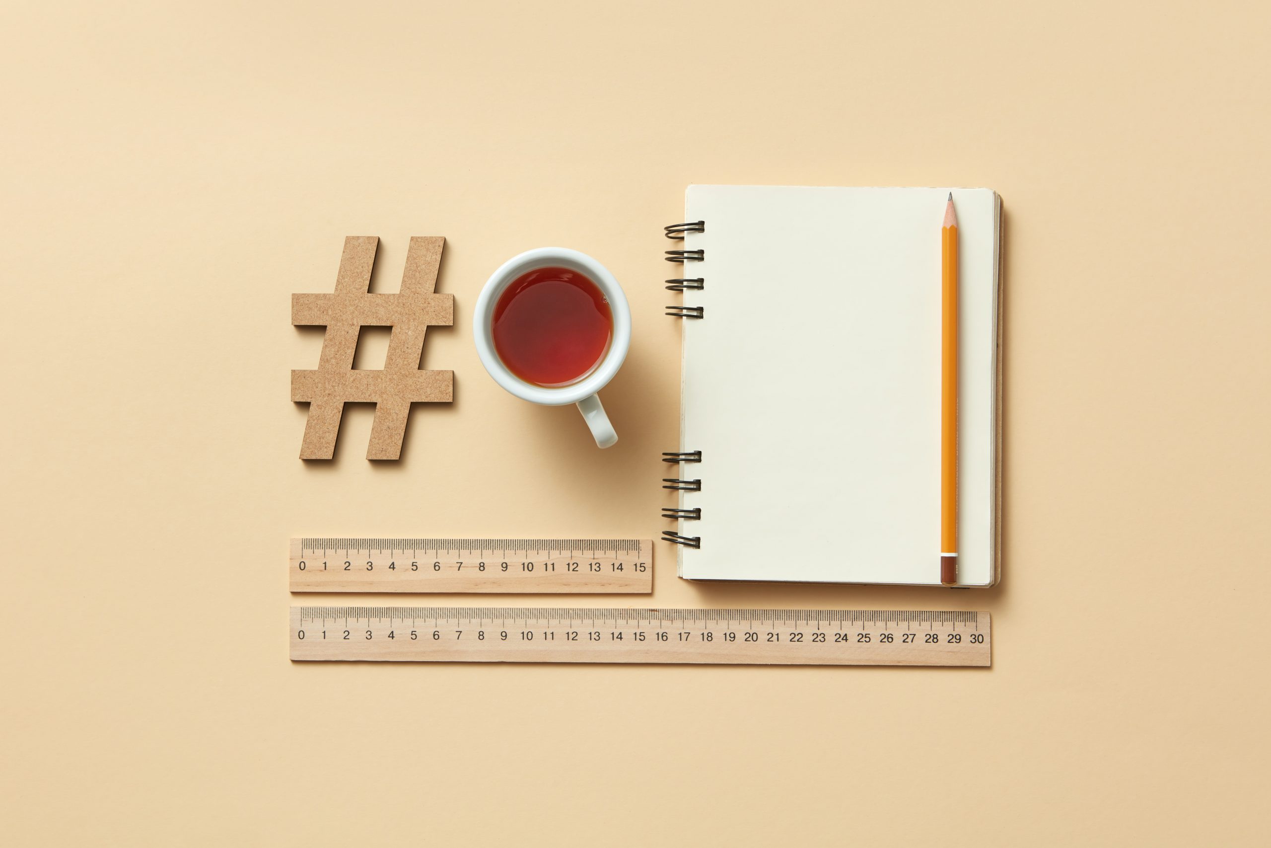 A flat lay image featuring a notebook, pencil, cup of tea, wooden hashtag symbol, and two wooden rulers on a beige background, suggesting a workspace or planning setup for a social media planner | plannthat.com
