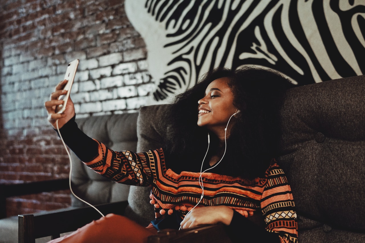 A woman with curly hair, wearing headphones and a colorful sweater, smiles while taking a selfie on her smartphone in a cozy room with a brick wall, using her Instagram scheduler | plannthat.com