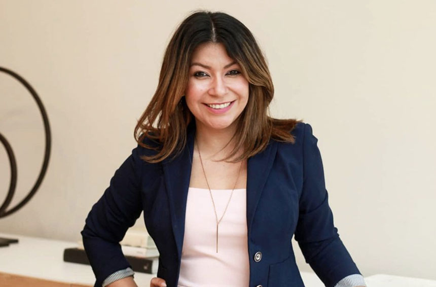 A woman with shoulder-length brown hair smiles confidently, wearing a navy blazer over a light pink top, standing with one hand on her hip against a neutral background, exuding professionalism and approachability, ideal for a social media planner | plannthat.com A woman with shoulder-length brown hair smiles confidently, wearing a navy blazer over a light pink top, standing with one hand on her hip against a neutral background, exuding professionalism and approachability, ideal for a social media planner | plannthat.com