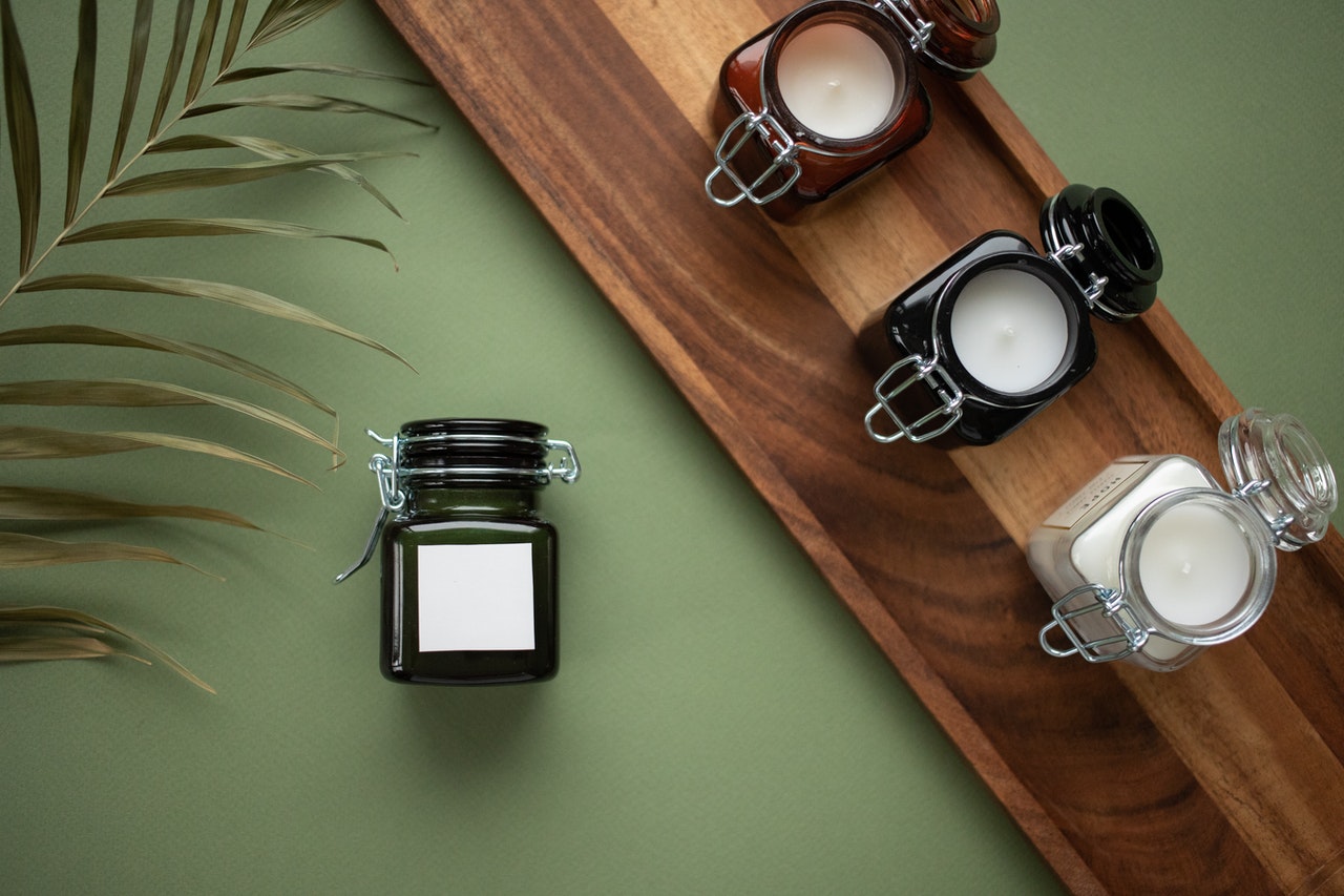A wooden tray with four candles in glass jars on a green background, accompanied by a green jar and a palm leaf, perfect for a serene Instagram scheduler post | plannthat.com