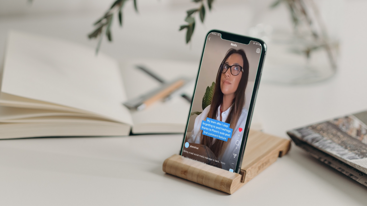 Smartphone on stand displaying a social media video captioned "My team after I stop forgetting to post hashtags thanks to Plann's auto post." Background includes an open notebook with a pen and pencil, and a magazine on a desk | Social media scheduler | plannthat.com