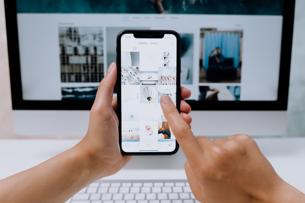 A person uses a smartphone to browse an Instagram profile, with a computer monitor displaying a similar interface in the background, suggesting multitasking and social media engagement in a modern, tech-savvy environment, ideal for a social media planner | plannthat.com