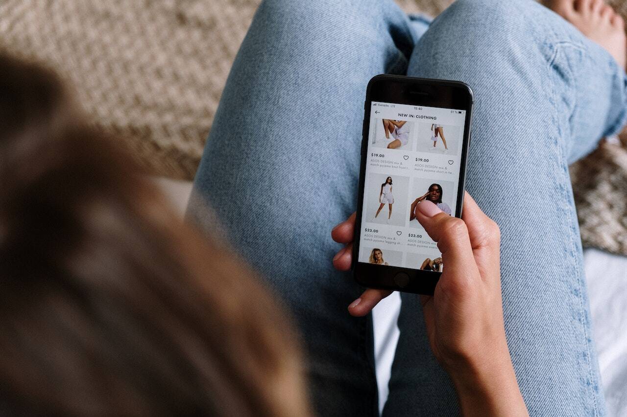 Person in blue jeans browsing an online clothing store on a smartphone, viewing various dresses and outfits with prices, using a social media scheduler | plannthat.com