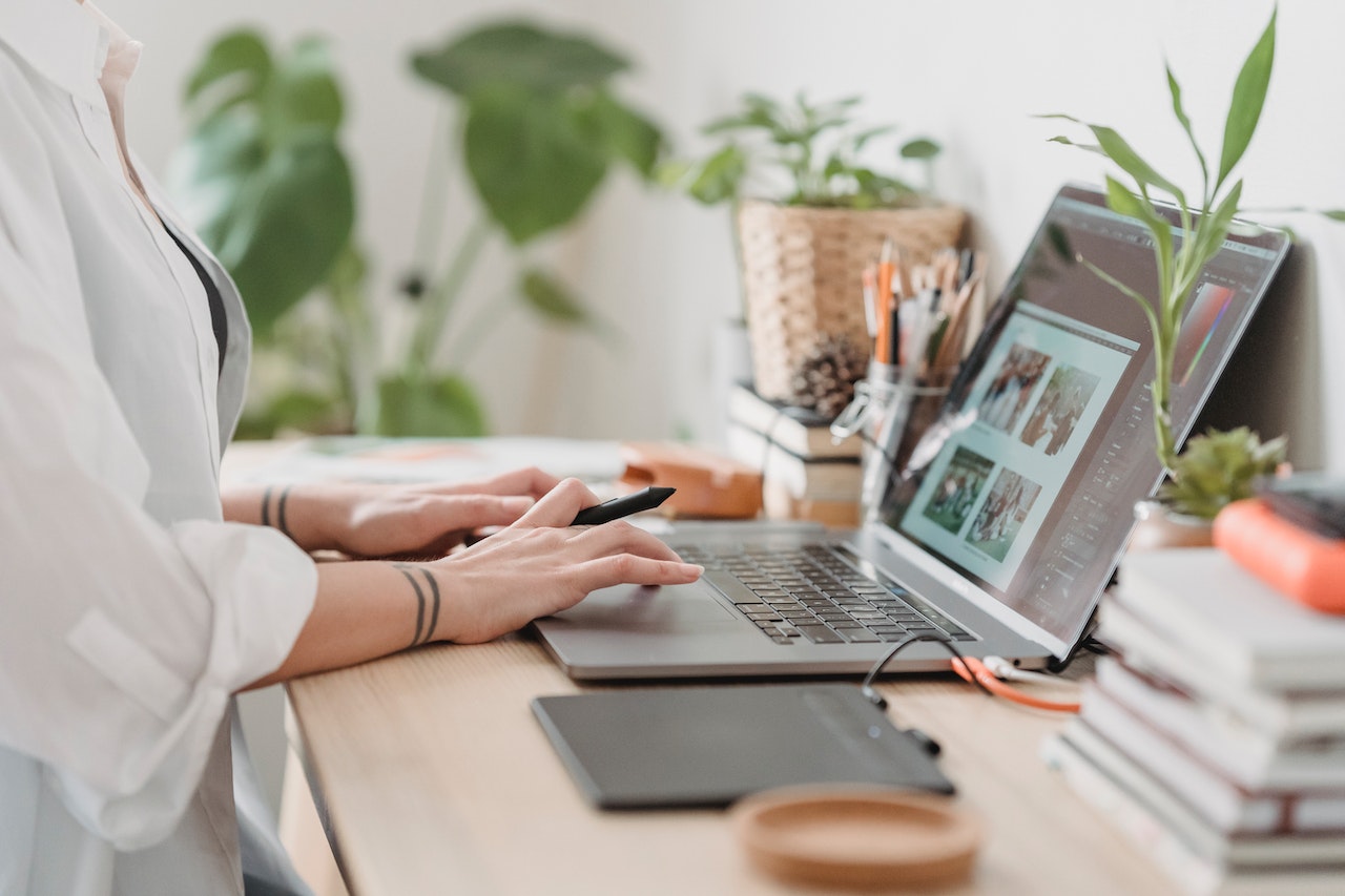 Person working on a laptop at a desk with plants and stationery, using a stylus and graphic tablet, planning social media content | plannthat.com