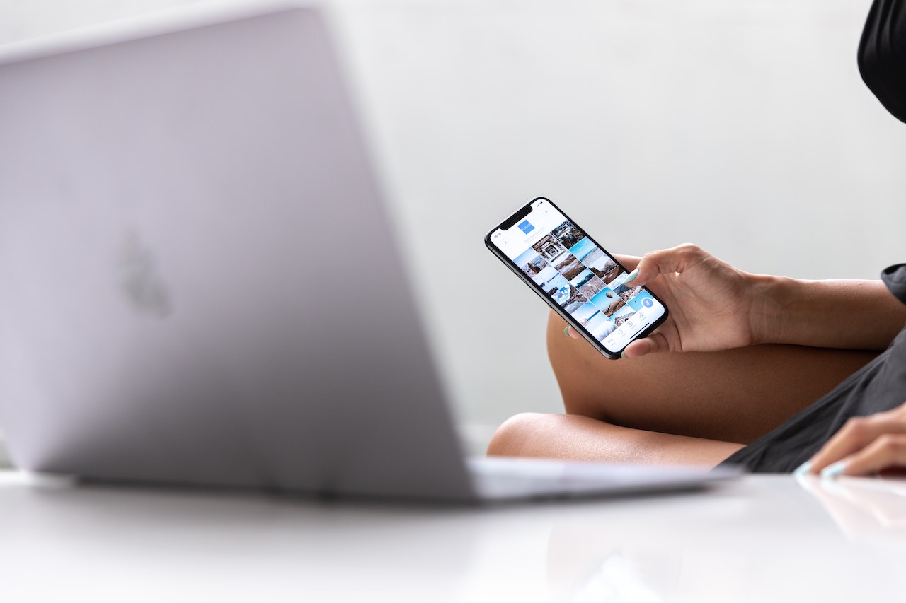 Person using a smartphone to browse photos, sitting next to a laptop on a white table. The focus is on the phone's screen displaying a photo gallery, possibly using a social media scheduler | plannthat.com