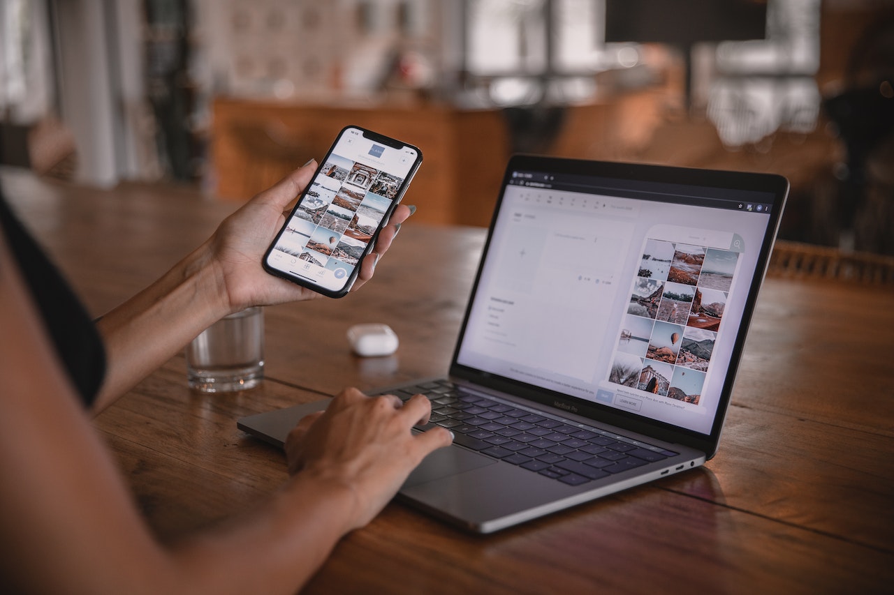 Person using a smartphone and laptop at a wooden desk, viewing photo galleries on both devices, with a glass of water and wireless earbuds nearby, illustrating the use of a social media scheduler | plannthat.com