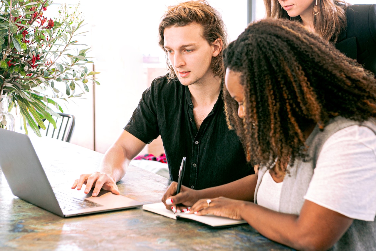 Three people collaborating at a table with a laptop and notebook, focused on work, with a vase of flowers in the background, using a social media scheduler | plannthat.com