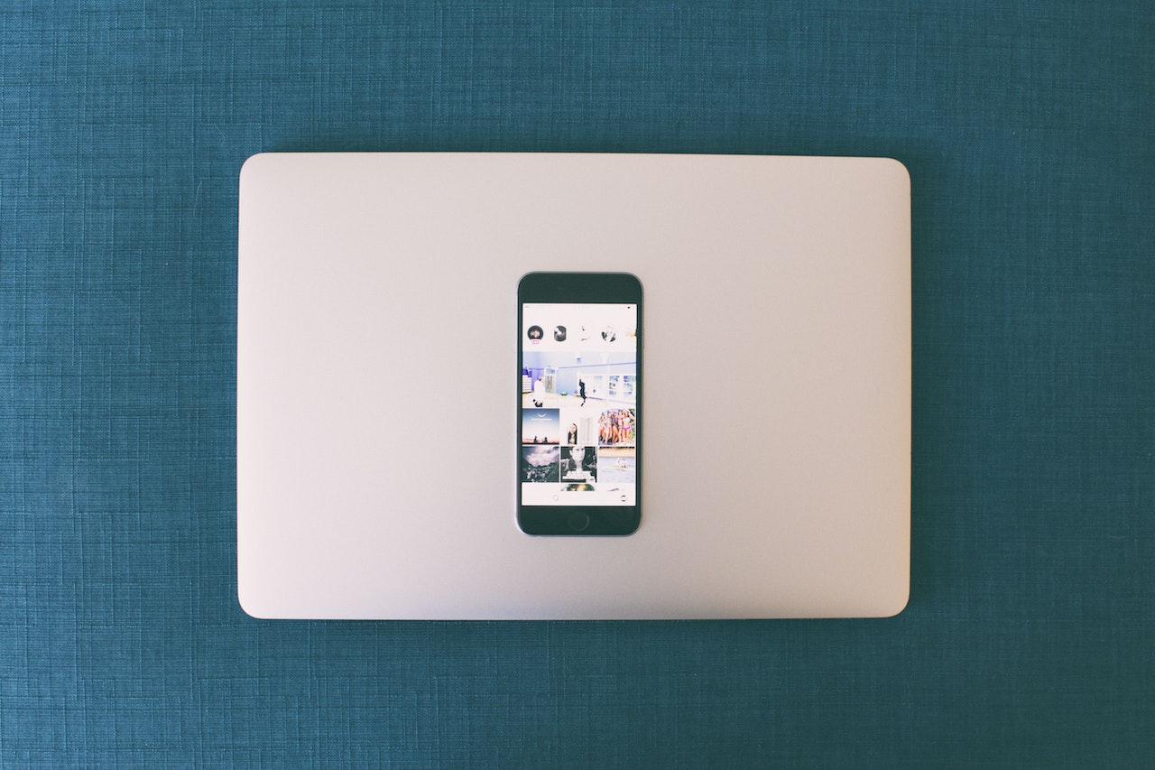 A smartphone displaying a social media feed on top of a closed laptop, both on a blue fabric surface, showcasing the integration of mobile and desktop technology in a minimalist setting with a focus on a social media scheduler | plannthat.com