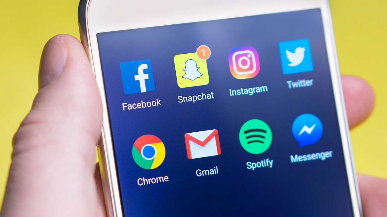 A close-up of a smartphone screen displaying various social media and app icons, including Facebook, Snapchat, Instagram, Twitter, Chrome, Gmail, Spotify, and Messenger, held by a hand against a yellow background, ideal for a social media scheduler | plannthat.com