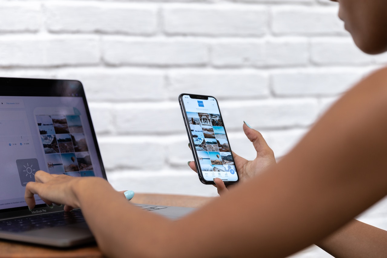 Person using a smartphone and a laptop simultaneously, browsing through photos on both devices against a white brick wall background, showcasing the use of a social media scheduler | plannthat.com