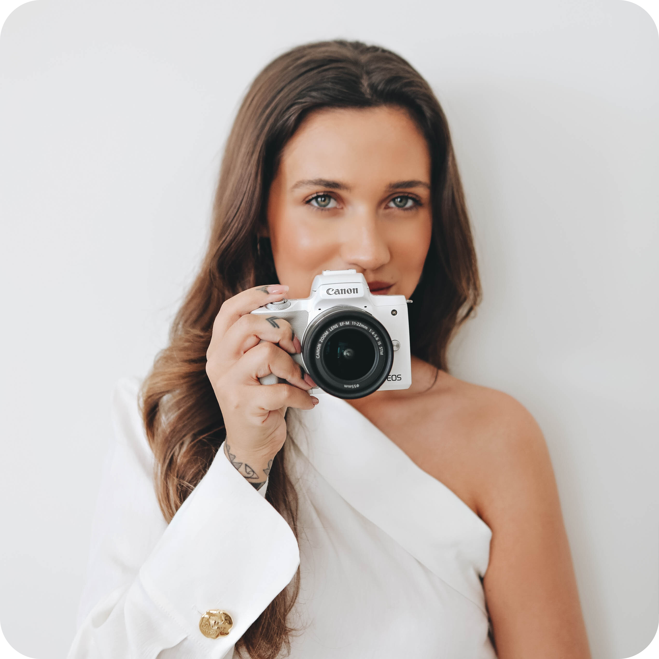 Woman in a white off-shoulder top holding a Canon EOS camera, looking directly at the lens, against a plain white background, perfect for a social media planner | plannthat.com Woman in a white off-shoulder top holding a Canon EOS camera, looking directly at the lens, against a plain white background, perfect for a social media planner | plannthat.com