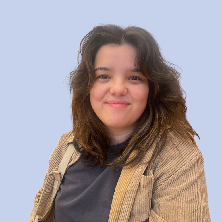 A person with shoulder-length dark hair, wearing a black shirt and beige jacket, smiles at the camera against a light blue background, appearing relaxed and approachable with a friendly expression, ideal for a social media planner | plannthat.com A person with shoulder-length dark hair, wearing a black shirt and beige jacket, smiles at the camera against a light blue background, appearing relaxed and approachable with a friendly expression, ideal for a social media planner | plannthat.com